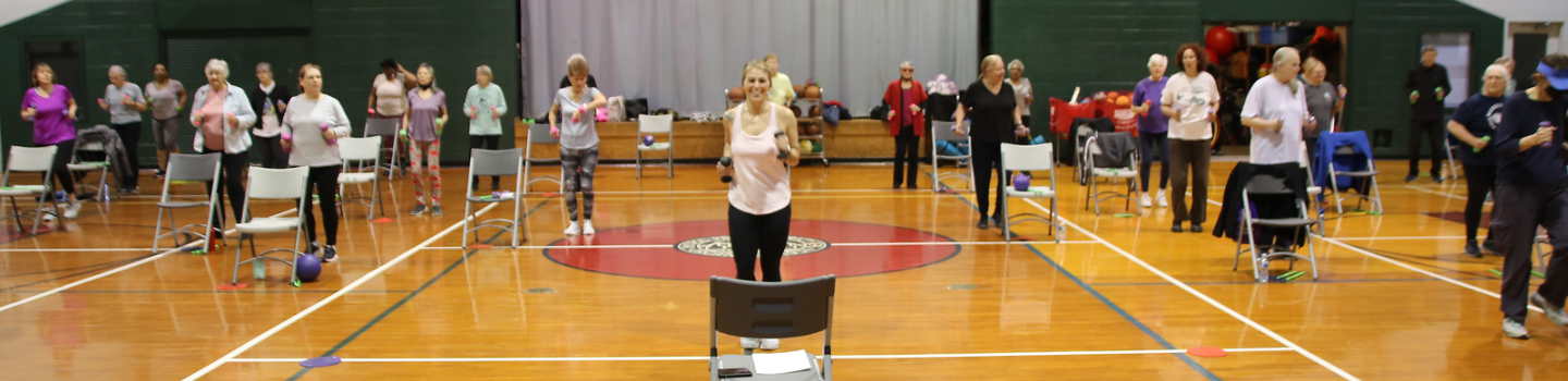 Seniors doing chair exercises in a gym. An instructor leads the class.